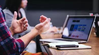 A person sitting in front of their laptop and an open book gestures as if in discussion with their co-workers who are in the background