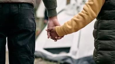 Taken from behind as an adult holds a child's hand. Together they face an out-of-focus tent in the background