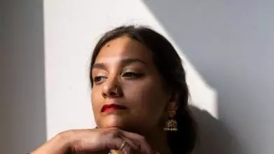Dr. Shyama Vermeersch is wearing striking red lipstick and is looking off to the side. She is also wearing a green scarf and has dark brown hair tied back.