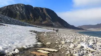 Three people walk across a beach in the artic. There is snow on the left and a moutain in the background.