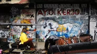 teenager in yellow t-shirt drives a horse drawn carriage down a street in indonesia