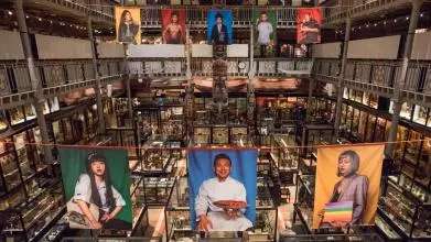 Panoramic view of the Performing Tibetan Identities exhibition inside the Pitt Rivers Museum. Large, photographic banners displaying people from the Tibetan community in in different styles of dress hang over the museum displays.