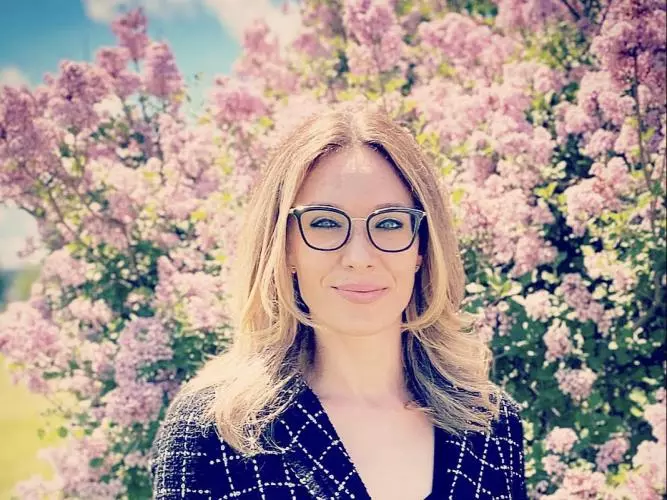 Celeste has long blonde hair, is wearing glasses and smiling into the camera