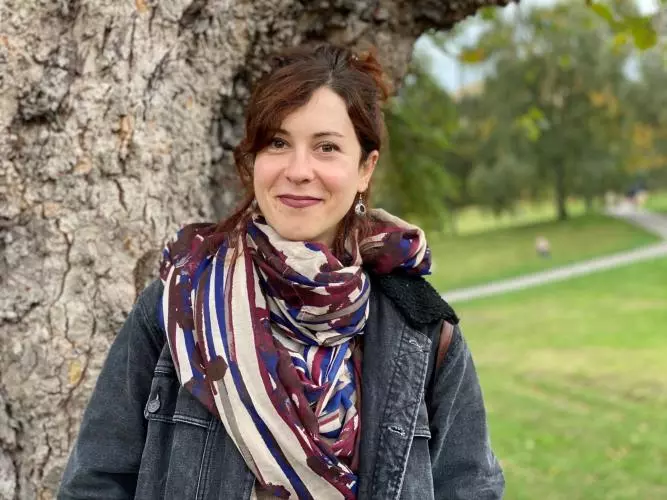 Dr Valdivia has long brown hair which she is wearing tied up. She is standing by a tree wearing a patterned cream, purple and blue scarf.