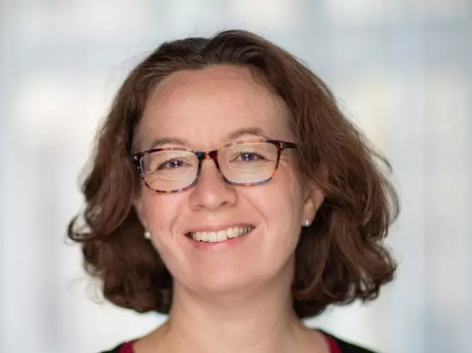 Dr Jones has short, curly brown hair. She is wearing dark glasses, a red dress and black cardigan and is smiling. 