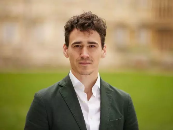 Dr Stephany is standing in front of a large patch of grass. He has short brown hair and is wearing a white shirt and grey blazer. 