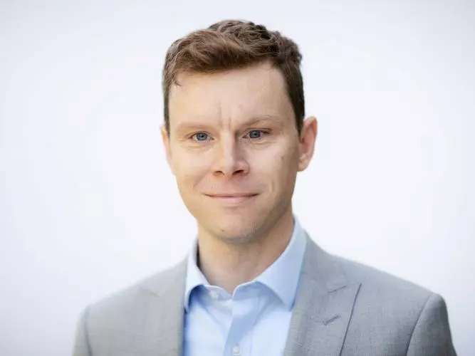 Professor Greg Taylor has short brown hair. He is wearing a light blue shirt and grey blazer. 