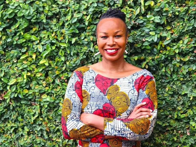 Dr Ogude-Chambert has dark braided hair and wearing a white top with patterns of bright red and yellow shapes. She is smiling and standing with her arms folded in front of some foliage.