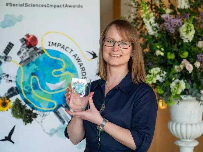 Dr Jennifer Seddon smiles and holds her prize trophy. In the background is the Impact Awards branding and a flower arrangement.