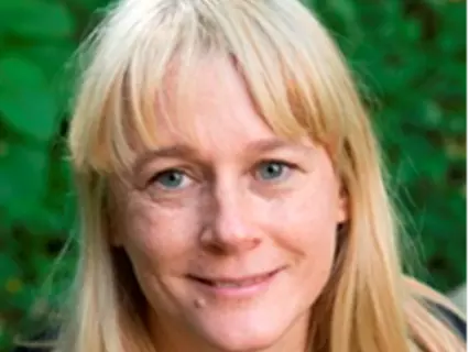 Dr Dingwall has long blonde hair with a fringe and blue eyes. She is standing in front of some green foliage and smiling.