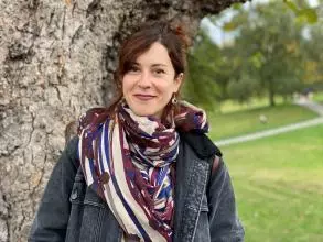 Dr Valdivia has long brown hair which she is wearing tied up. She is standing by a tree wearing a patterned cream, purple and blue scarf.