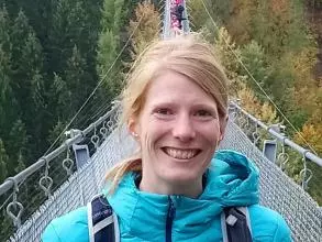 Dr Styring is standing on a suspension bridge, smiling. She has blonde hair which is tied back, and is wearing a bright blue coat with a black rucksack. 