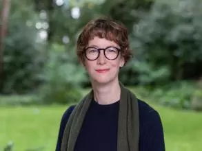 Dr Hamilton has short auburn hair and is wearing dark-framed round glasses. She is wearing a navy jumper and dark green scarf, standing in a garden.