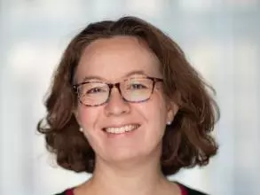 Dr Jones has short, curly brown hair. She is wearing dark glasses, a red dress and black cardigan and is smiling. 