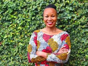 Dr Ogude-Chambert has dark braided hair and wearing a white top with patterns of bright red and yellow shapes. She is smiling and standing with her arms folded in front of some foliage.