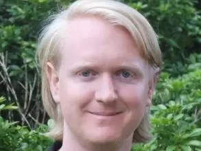 Dr Wainwright has long blonde hair and is wearing a black t-shirt and blazer. He is standing in front of foliage and smiling. 