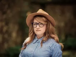 Dr Gemma Newlands has long auburn wavy hair and large, blue-rimmed glasses. She is wearing a straw hat and a blue and white striped shirt, looking directly at the camera. 
