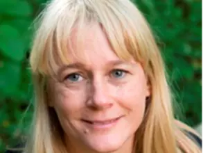 Dr Dingwall has long blonde hair with a fringe and blue eyes. She is standing in front of some green foliage and smiling.