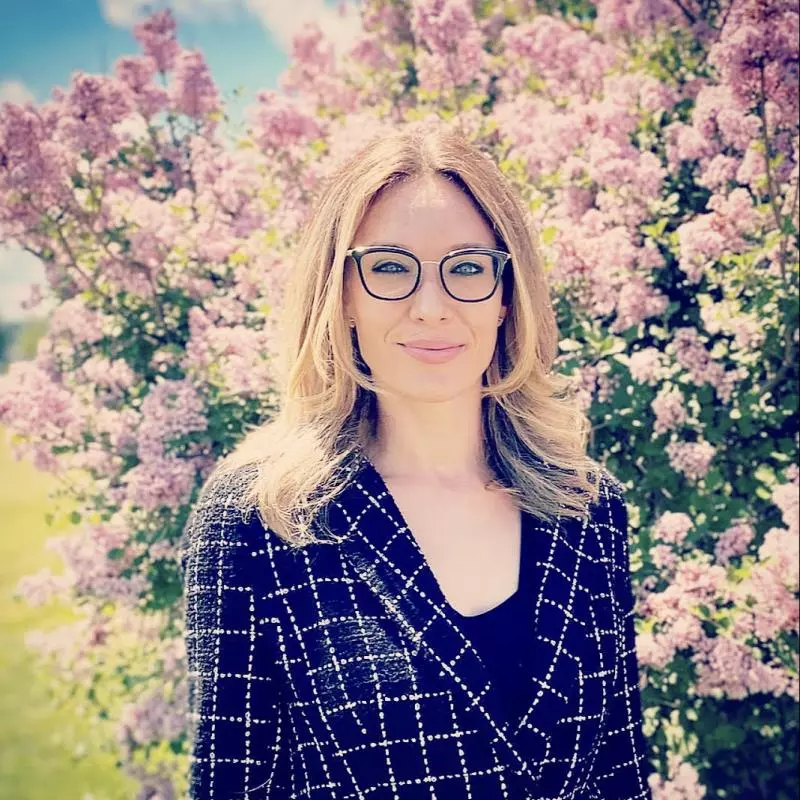 Celeste has long blonde hair, is wearing glasses and smiling into the camera