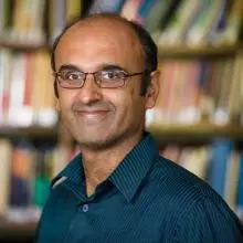 Portrait photo of Professor Yadvinder Malhi, smiling against a blurred background of shelves full of books