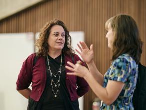 Two women speaking to each other, one blurred in the foreground is gesturing and the other is listening