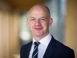 Paul Chapman is wearing a navy suit, white shirt and striped tie. He is looking directly at the camera and smiling. 