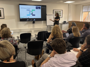 A room of seated people listening to Prof Rebecca Williams present the AIEOU Hub