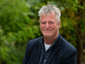 Professor Steve Strand has short grey hair and is wearing a navy blazer over a blue shirt and white top. He is outside in front of some green foliage.