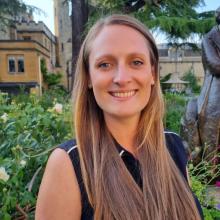 Naomi is standing in a garden smiling at the camera. She has long blonde hair and is wearing a navy blouse.