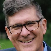White man with dark rimmed glasses and dark hair smiles at the camera