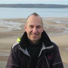 White man smiles at camera with a beach and sea background