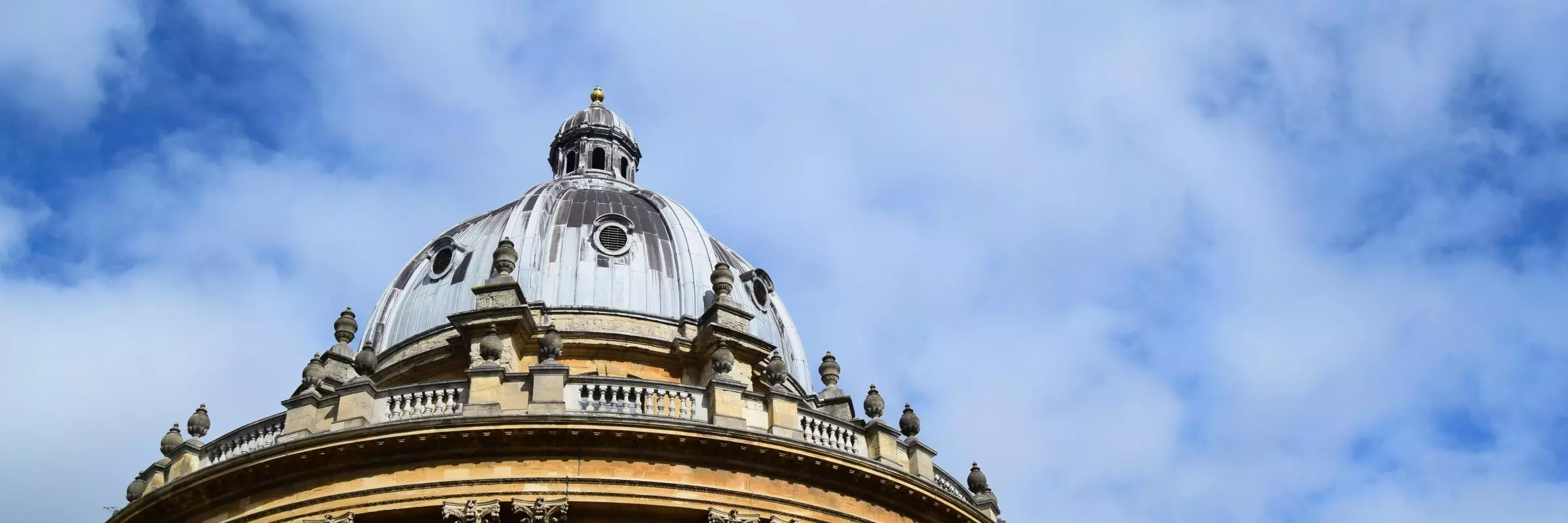 A view of the circular Radcliffe Camera library showing the blue-grey leaded roof and yellow standstone walls against a bright blue sky with whispy clouds