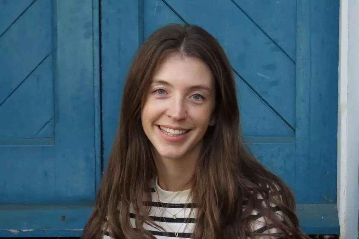 Dr Steele has long brown hair and smiles while sitting in front of a blue wooden door. She is wearing a white and navy striped shirt.