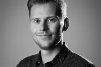 A black-and-white portrait of Dr Brindle with short hair and a trimmed beard, smiling slightly. He is wearing a dark, button-up shirt with a small dotted pattern