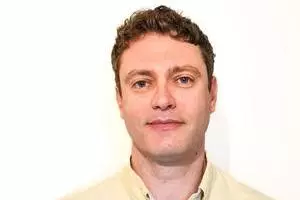 Dr Robins has short curly hair and is wearing a light yellow shirt against a plain white background