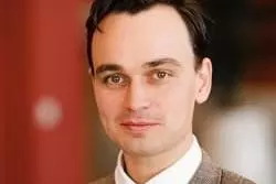 Dr Poore has dark hair and is wearing a brown suit jacket, white shirt, and tie, posing indoors.