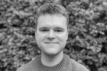 Black and white image of Dr Thomsen with short hair smiling, wearing a knitted sweater, standing in front of leafy bushes.